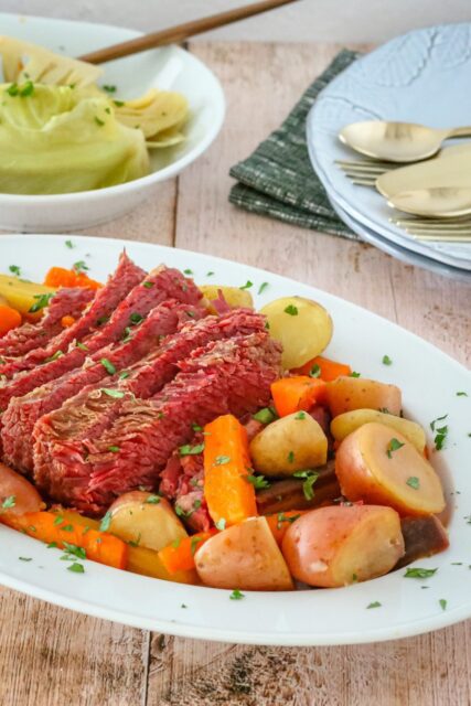 corned beef on a platter with potatoes, carrots, and cabbage on a plate in the background