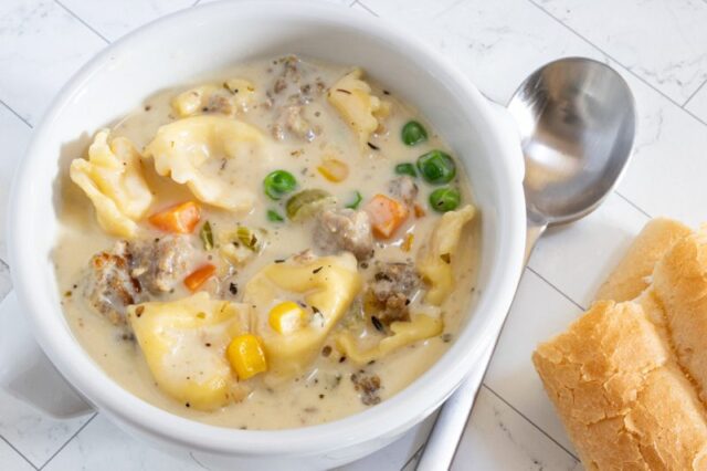 A bowl of sausage and tortellini soup with bread on the side.