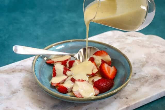 Cream being poured over sliced strawberries and bananas in a bowl.