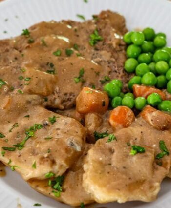 cube steak with mushrooms and gravy shown on a plate with potatoes, peas, and carrots
