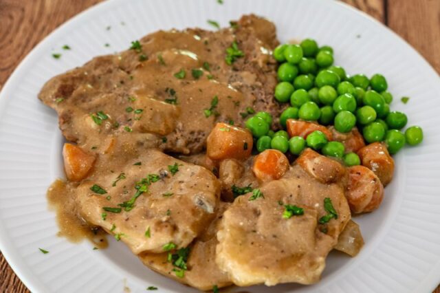 cube steak with mushrooms and gravy shown on a plate with potatoes, peas, and carrots