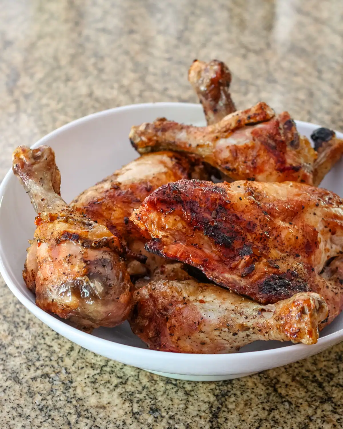 Juicy grilled chicken drumsticks in a white bowl on a granite countertop.