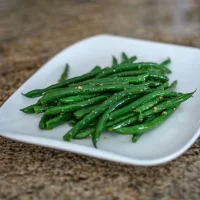 Steamed green beans seasoned with garlic on a white square plate.