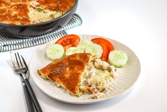 A ham and leek pie with puff pastry on a plate served with cucumbers and tomato wedges.