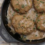 An iron skillet with hamburger steaks and gravy, with a garnish od sliced chives.