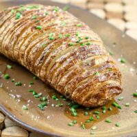 Golden-brown Hasselback potato on a serving platter with green garnishes.
