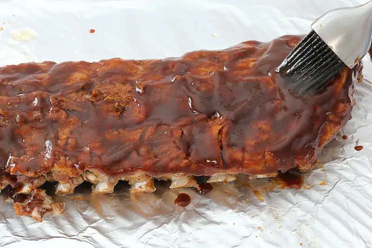 Baked rack of ribs covered in barbecue sauce on foil surface.
