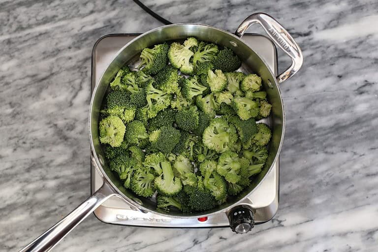 Steamed broccoli florets in a metal steamer basket on a marble countertop.