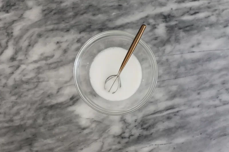 Whisk and milk mixture in a glass bowl on marble surface.