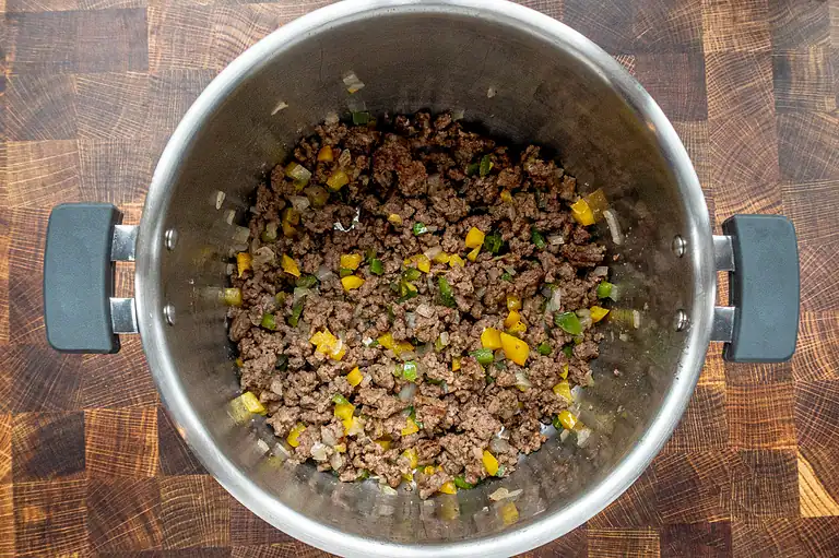 Ground beef cooked with diced onions, green and yellow bell peppers in a stainless steel skillet.