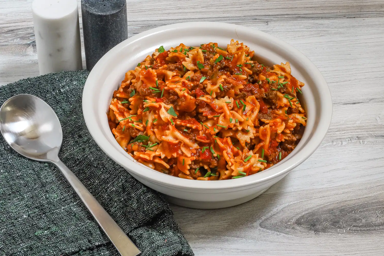 A serving bowl of farfalle pasta with ground beef and tomatoes, garnished with chives and parsley.