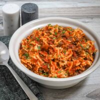 A serving bowl of farfalle pasta with ground beef and tomatoes, garnished with chives and parsley.