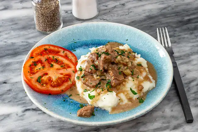 Instant Pot beef tips and gravy on mashed potatoes with tomatoes on the side.