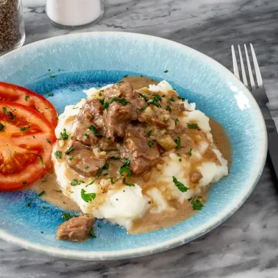 Sliced tomatoes served on a blue ceramic plate with beef stew and mashed potatoes garnished with herbs.