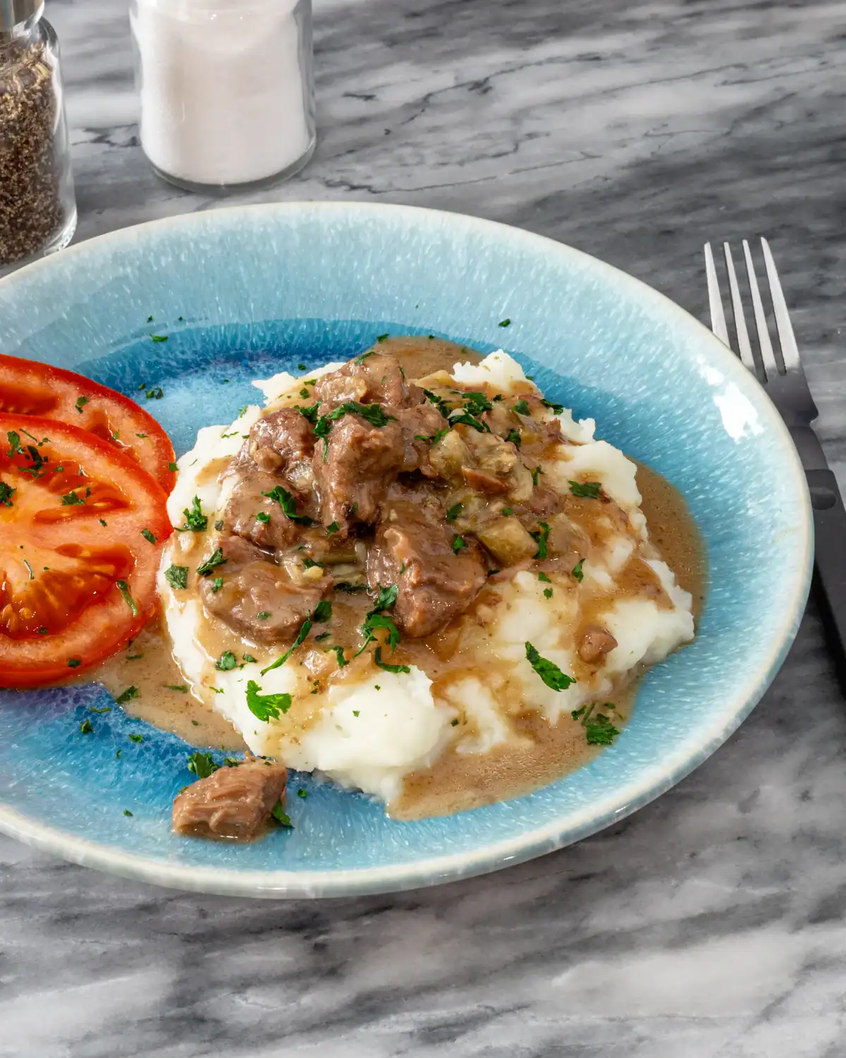 Sliced tomatoes served on a blue ceramic plate with beef stew and mashed potatoes garnished with herbs.