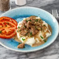 Creamy beef stew served over mashed potatoes with sliced tomatoes on the side.