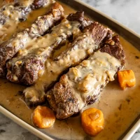 Beef short ribs topped with creamy mushroom gravy and carrots in a baking dish.