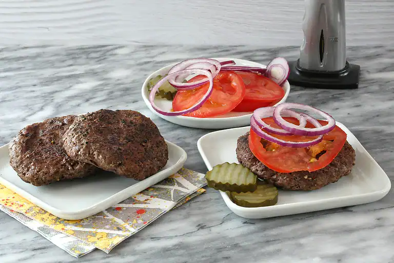 Grilled beef patties with tomato slices, onions, and pickles on white plates.