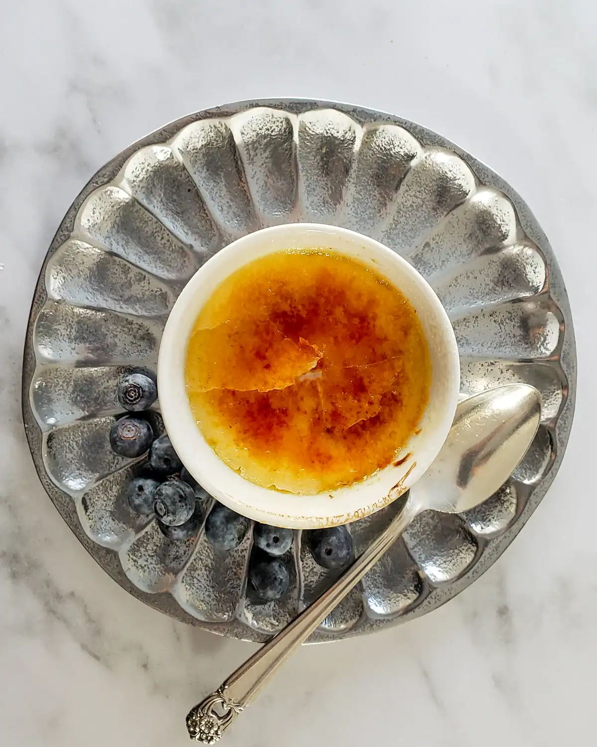 Creme brulee in a small white ramekin on a decorative silver plate with blueberries.