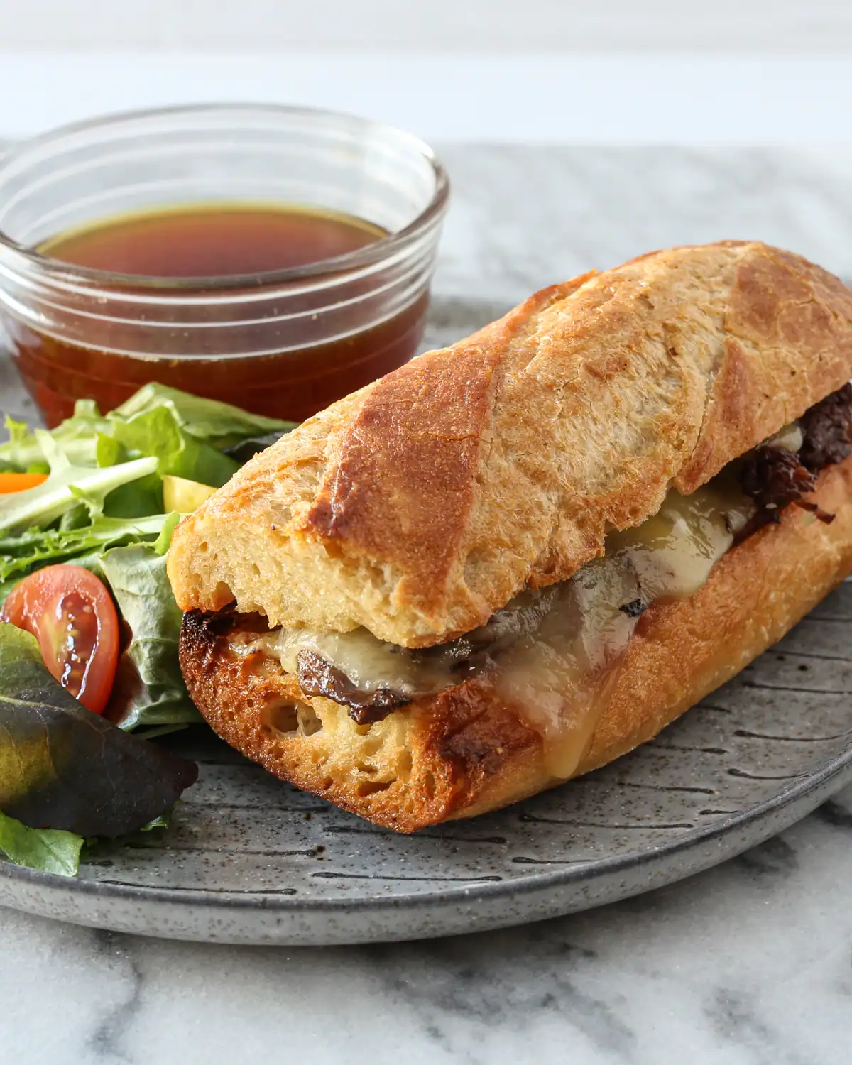 Crusty baguette sandwich with melted cheese and grilled meats on a gray plate.