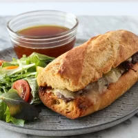French baguette sandwich with melted cheese and beef patty on side salad.