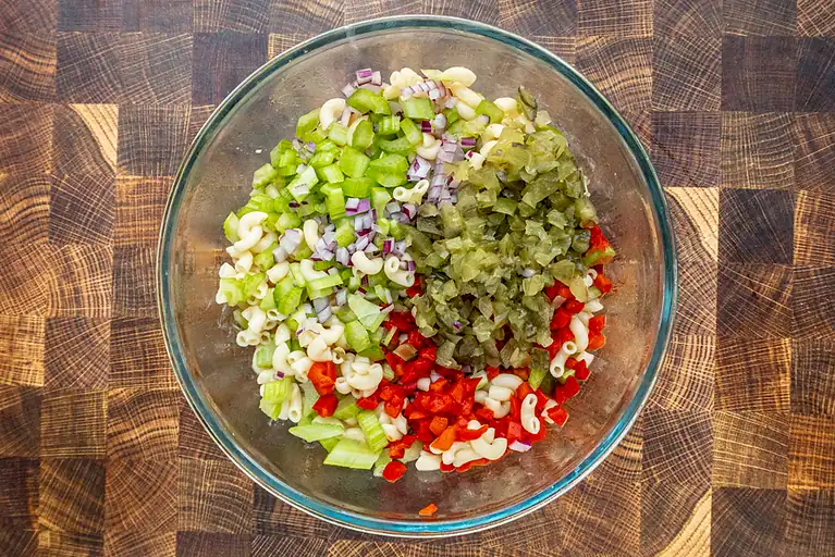 Diced celery, pickles, red bell peppers, and macaroni in a glass bowl.