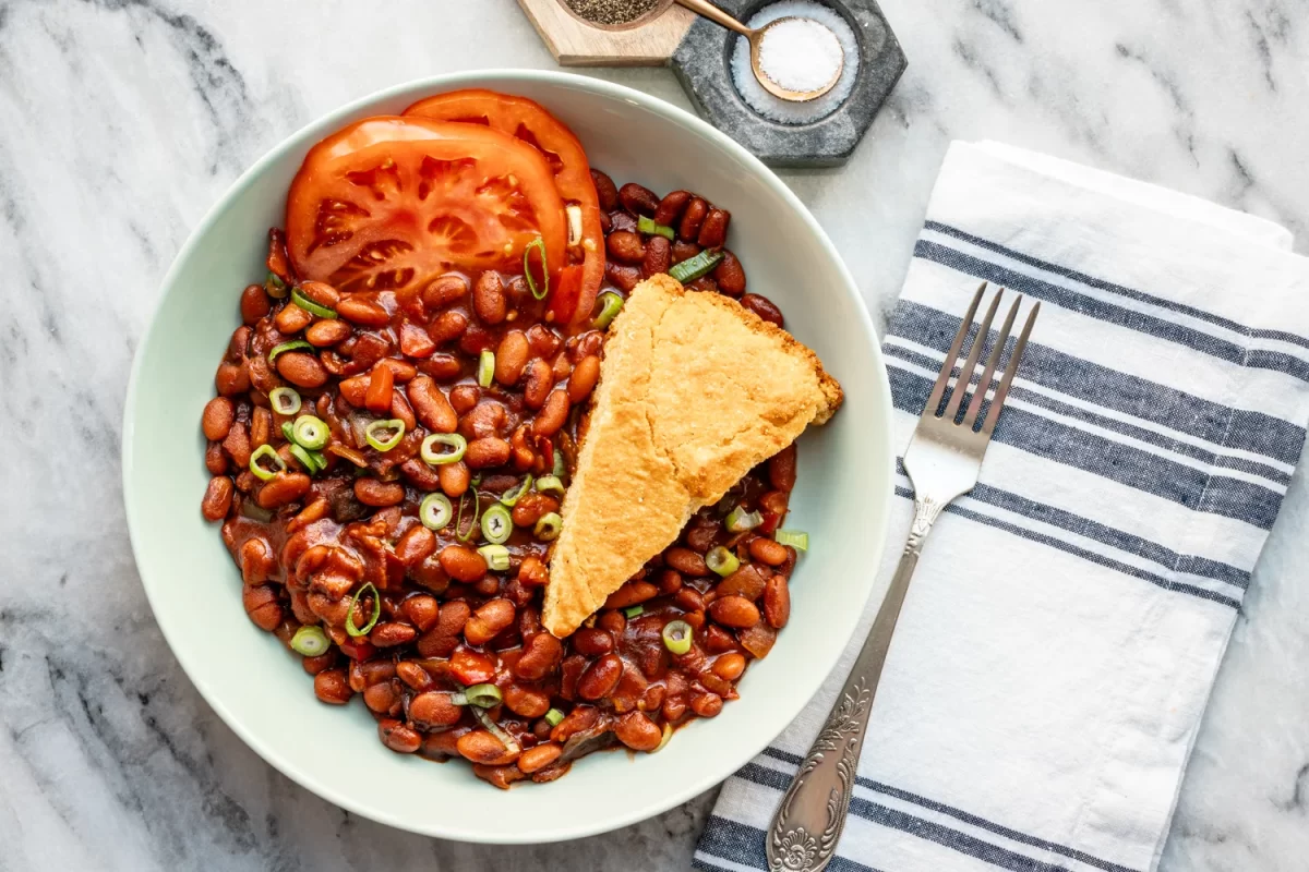 A serving dish with Instant Pot cooked pinto beans with a wedge of cornbread, tomato slices, and sliced green onions.