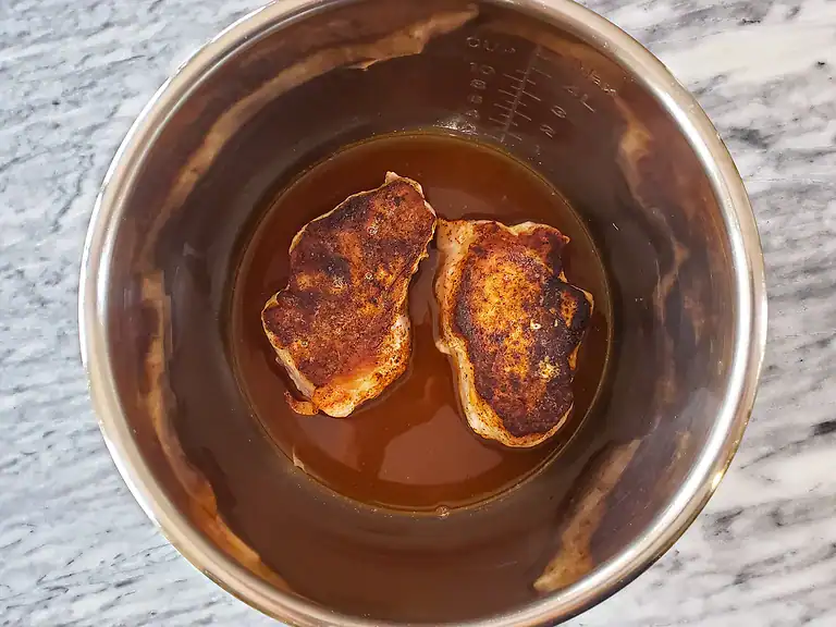 Fried pork chops cooking in a brown sauce inside a stainless steel cooking pot.