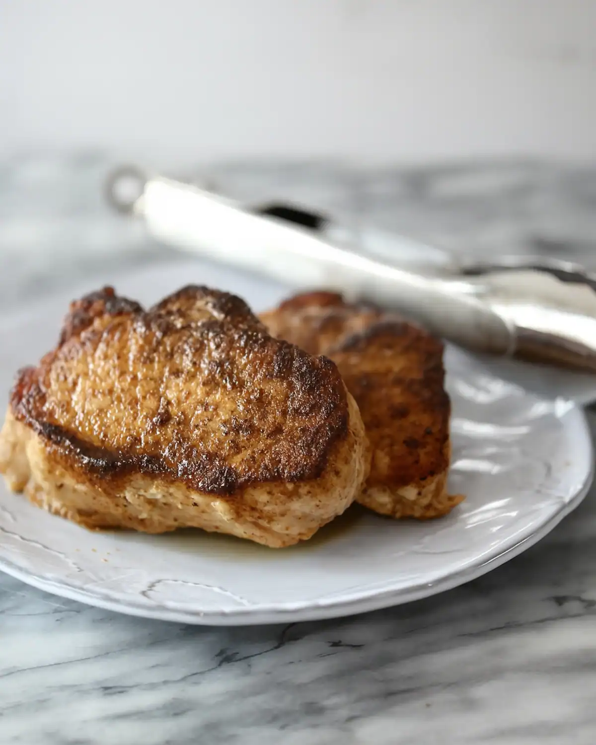 Grilled chicken thighs on a white plate with tongs nearby.