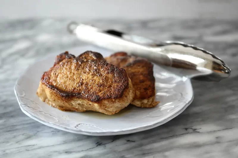 Crispy seared pork chop on white plate with tongs in background.