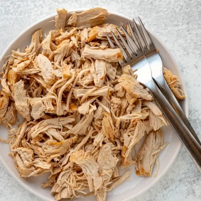 Shredded cooked chicken on a white plate with a fork and knife.