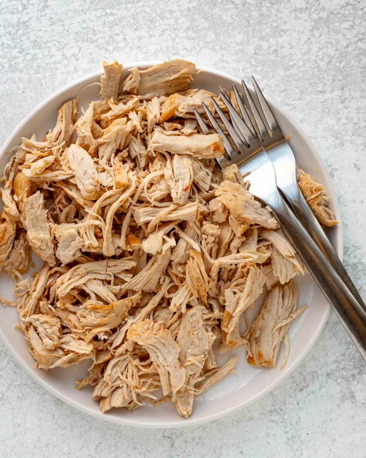 Shredded cooked chicken on a white plate with a fork and knife.