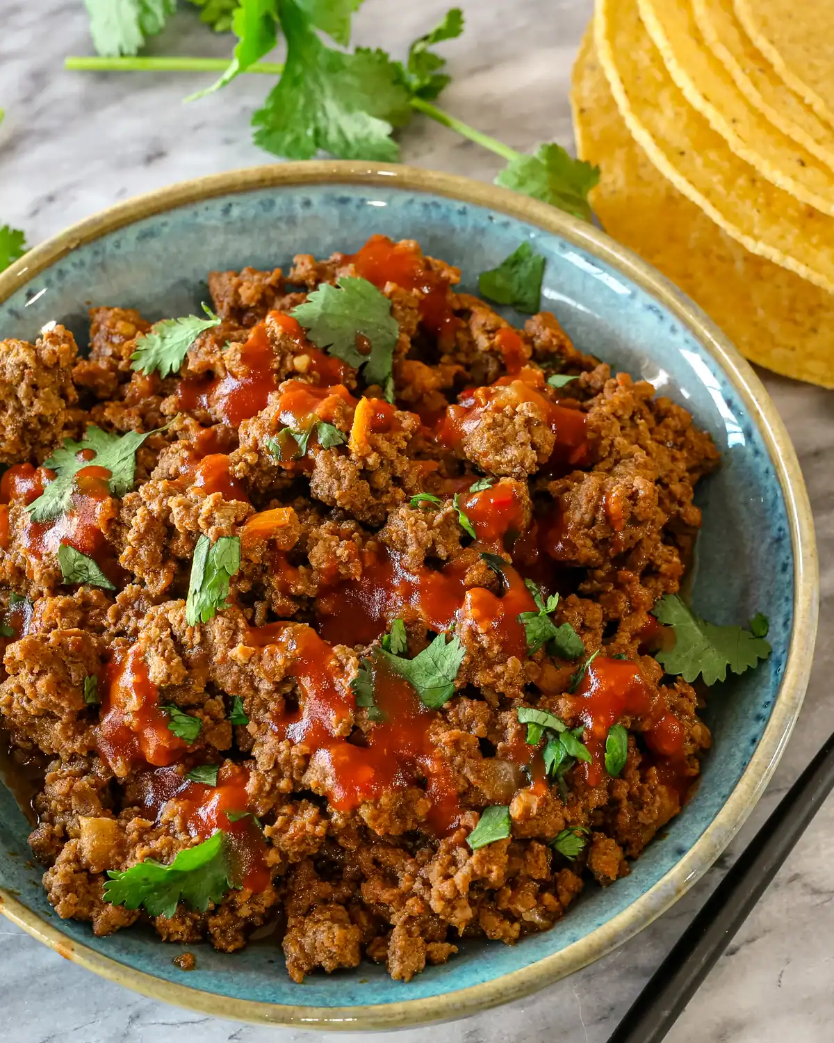 Ground beef chili in a blue bowl with cilantro garnishes and chili sauce.