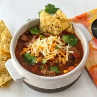Recommendations, chili, with shredded cheese, Garnished with cilantro and tortilla chips, Hearty beef chili with vegetables served in a white bowl.