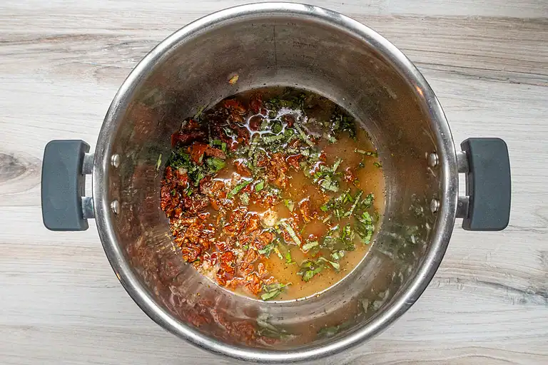 Sun-dried tomatoes, herbs, and broth in a stainless steel pot.