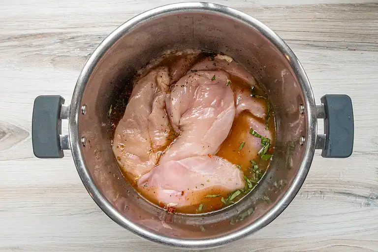 Raw chicken breasts marinating in a bowl with herbs and sauce.