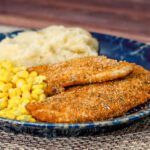 Oven fried chicken breasts on a plate with corn and mashed potatoes.
