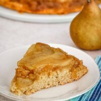 A slice of pear upside-down cake on a plate with a pear in the background.