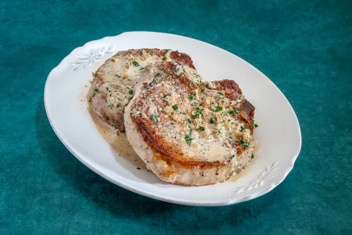 Pork chops are on a serving platter with a pan sauce and garnish of parsley.