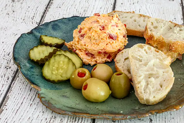 Pimiento cheese on a plate with olives, pickles, and baguette slices.