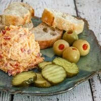 Pimiento cheese on a plate with olives, pickles, and baguette slices.