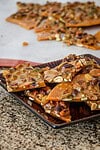 Butterscotch pecan brittle pieces with nuts on a black tray in front of a larger batch of brittle.