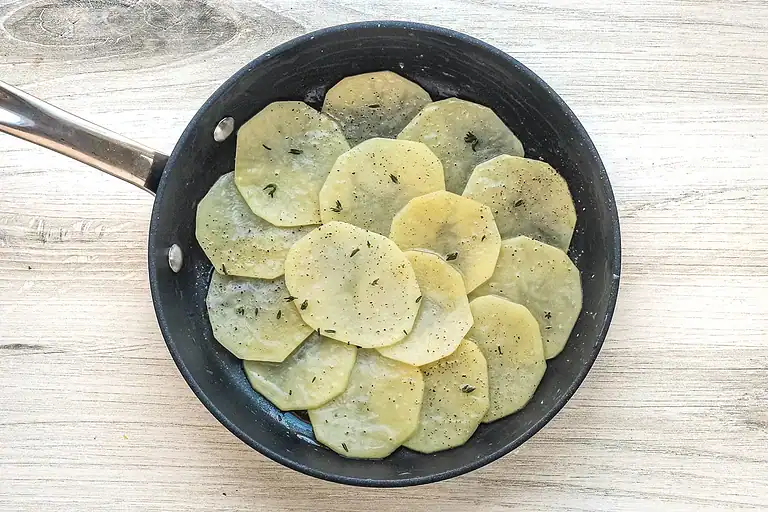Thinly sliced potatoes cooking in a skillet with herbs and black pepper.