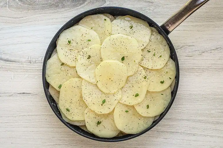 Thinly sliced potatoes seasoned with herbs in a frying pan.