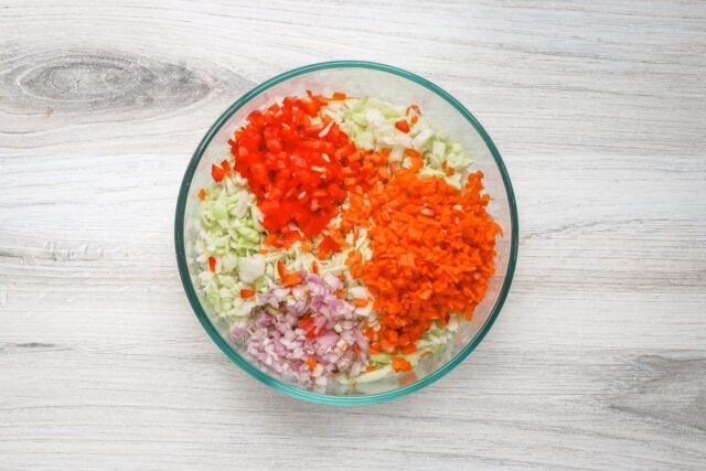 Chopped red bell peppers, onions, carrots, and celery in a glass bowl.