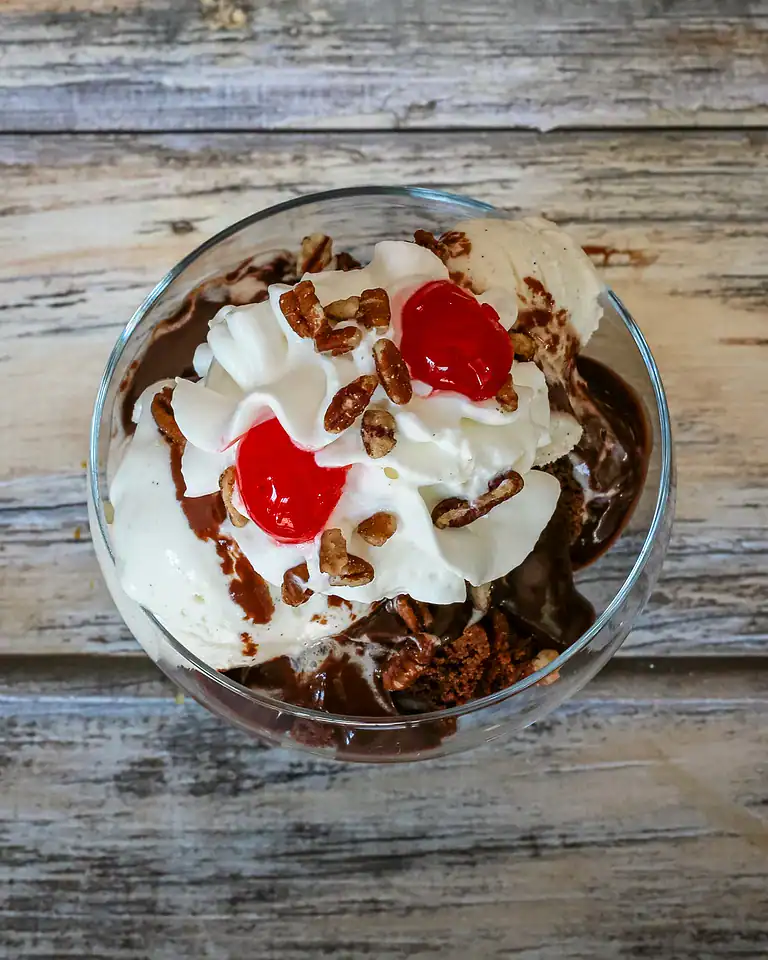 Rich chocolate ice cream topped with whipped cream, cherries, and chopped nuts in a glass dish.