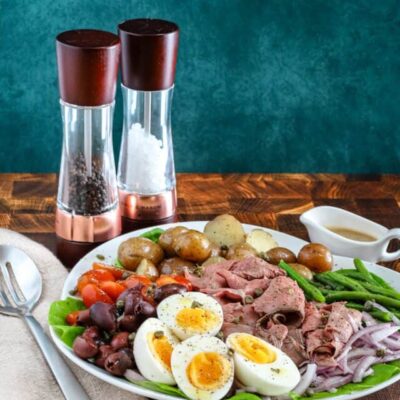 Roast beef Nicoise salad on a platter includes capers, eggs, deli sliced roast beef, tomatoes, olives, potatoes, onions, green beans, and a Dijon mustard dressing.