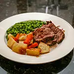 Roast beef with roasted potatoes, carrots, and green peas on white plate.