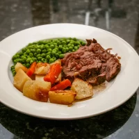 Roast beef with roasted potatoes, carrots, and green peas on white plate.