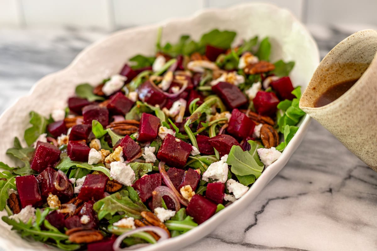 A serving dish of roasted beet salad with vinaigrette being drizzled.